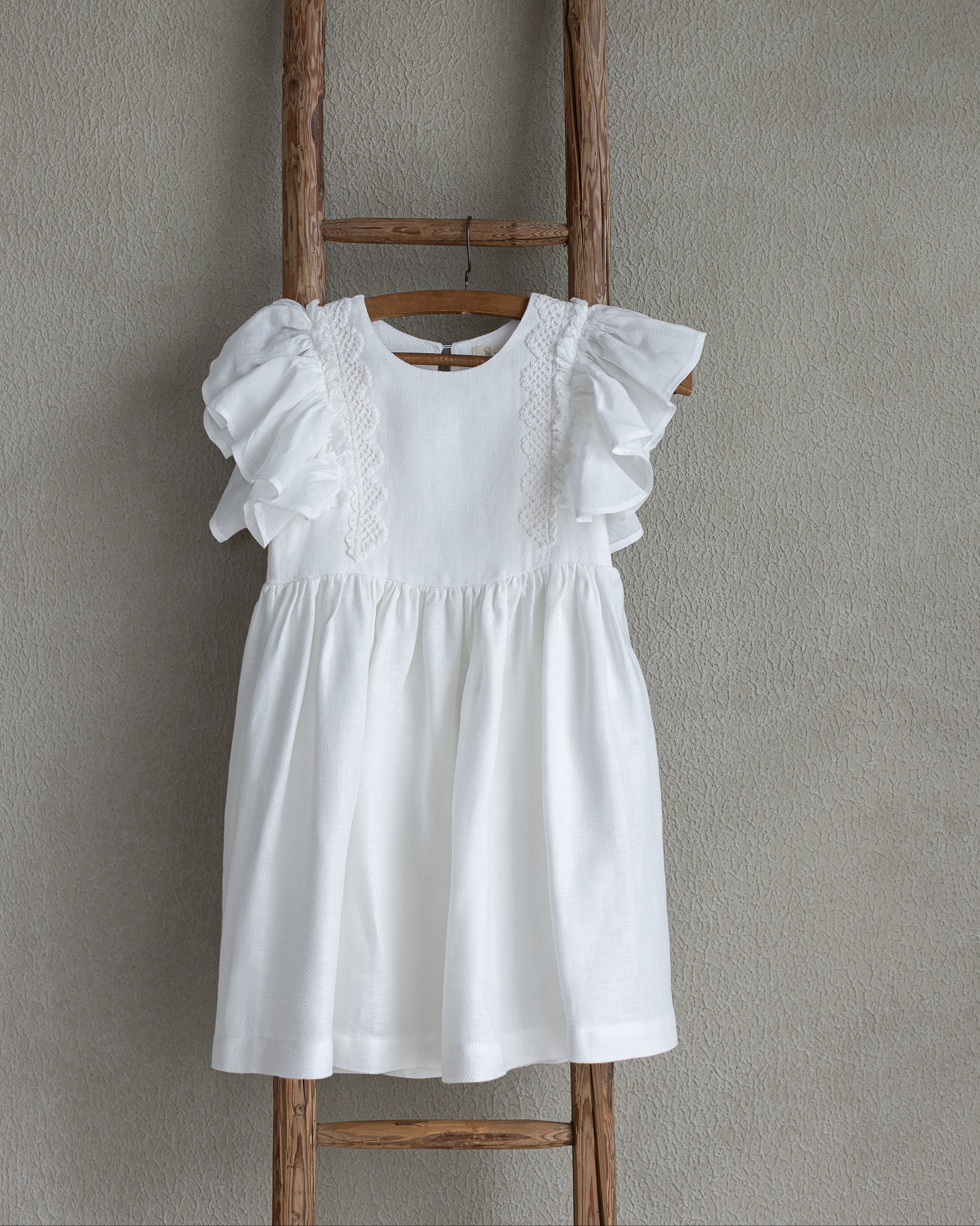 White linen dress with ruffled sleeves and crochet lace on a wooden ladder against a gray wall