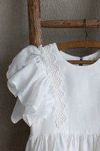 White linen dress with ruffled sleeves and crochet lace on a wooden ladder against a neutral wall.