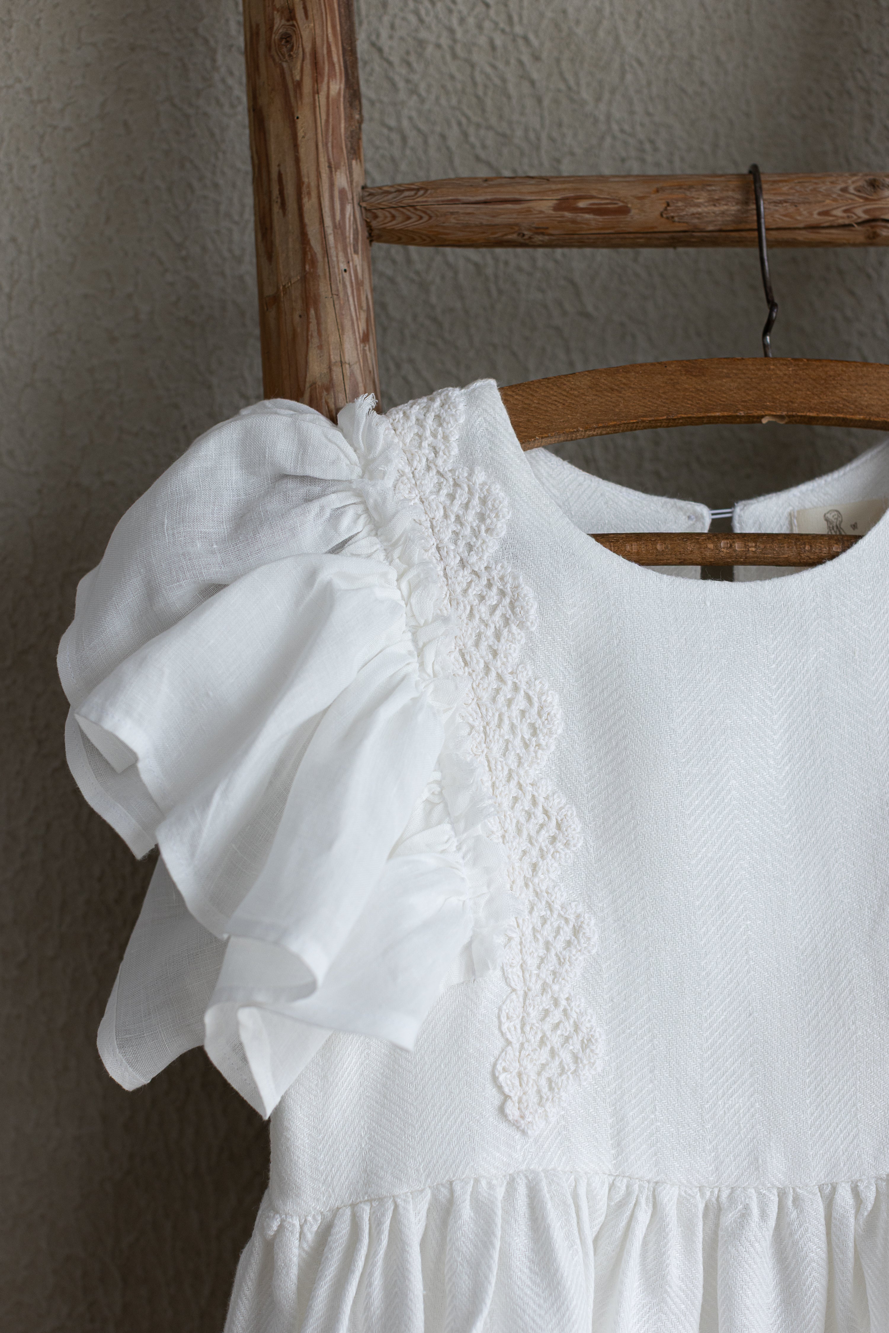White linen dress with ruffled sleeves and crochet lace on a wooden ladder against a neutral wall.