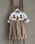 Children's linen dress with floral crochet motif on a wooden ladder against a plain wall.