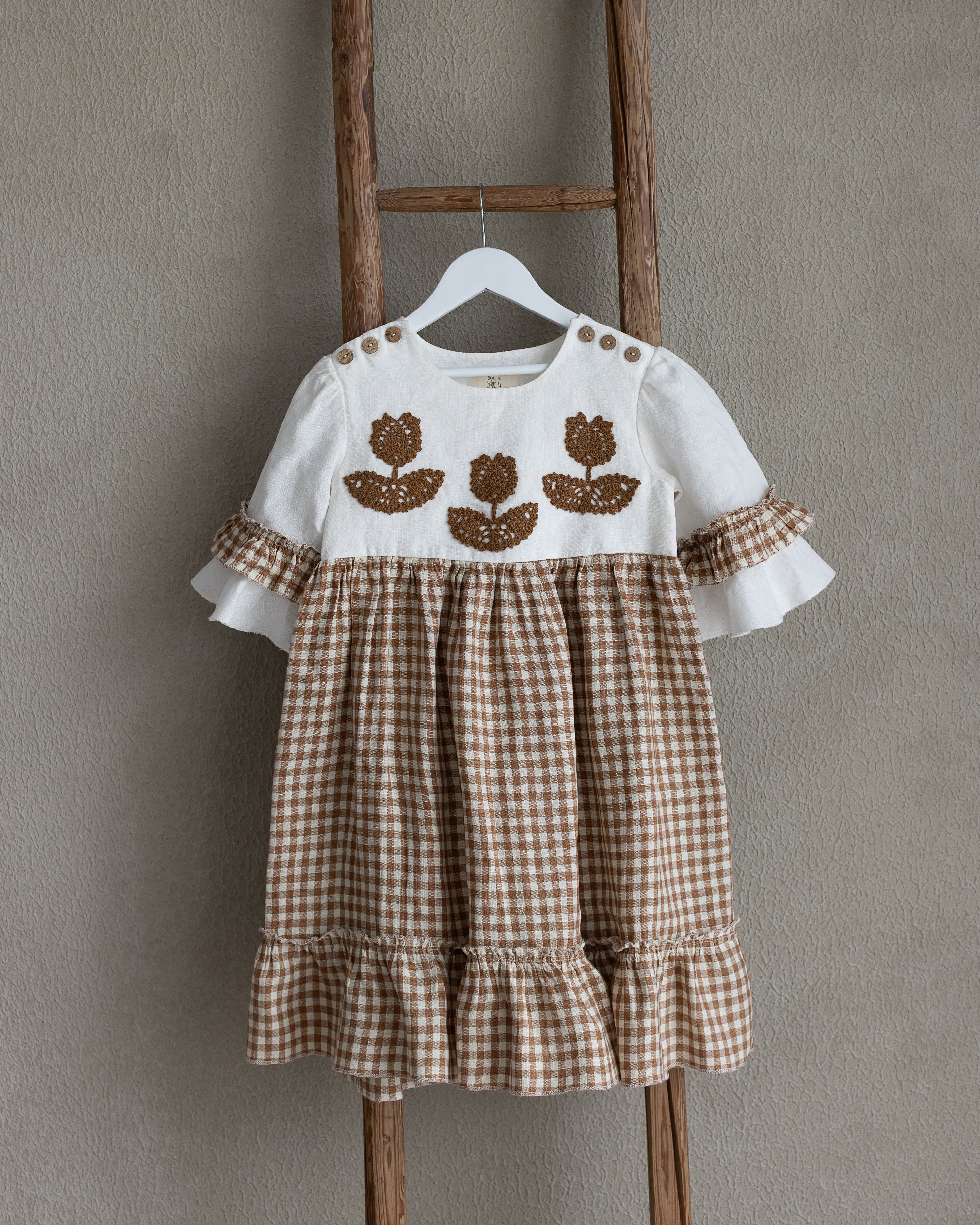 Children's linen dress with floral crochet motif on a wooden ladder against a plain wall.