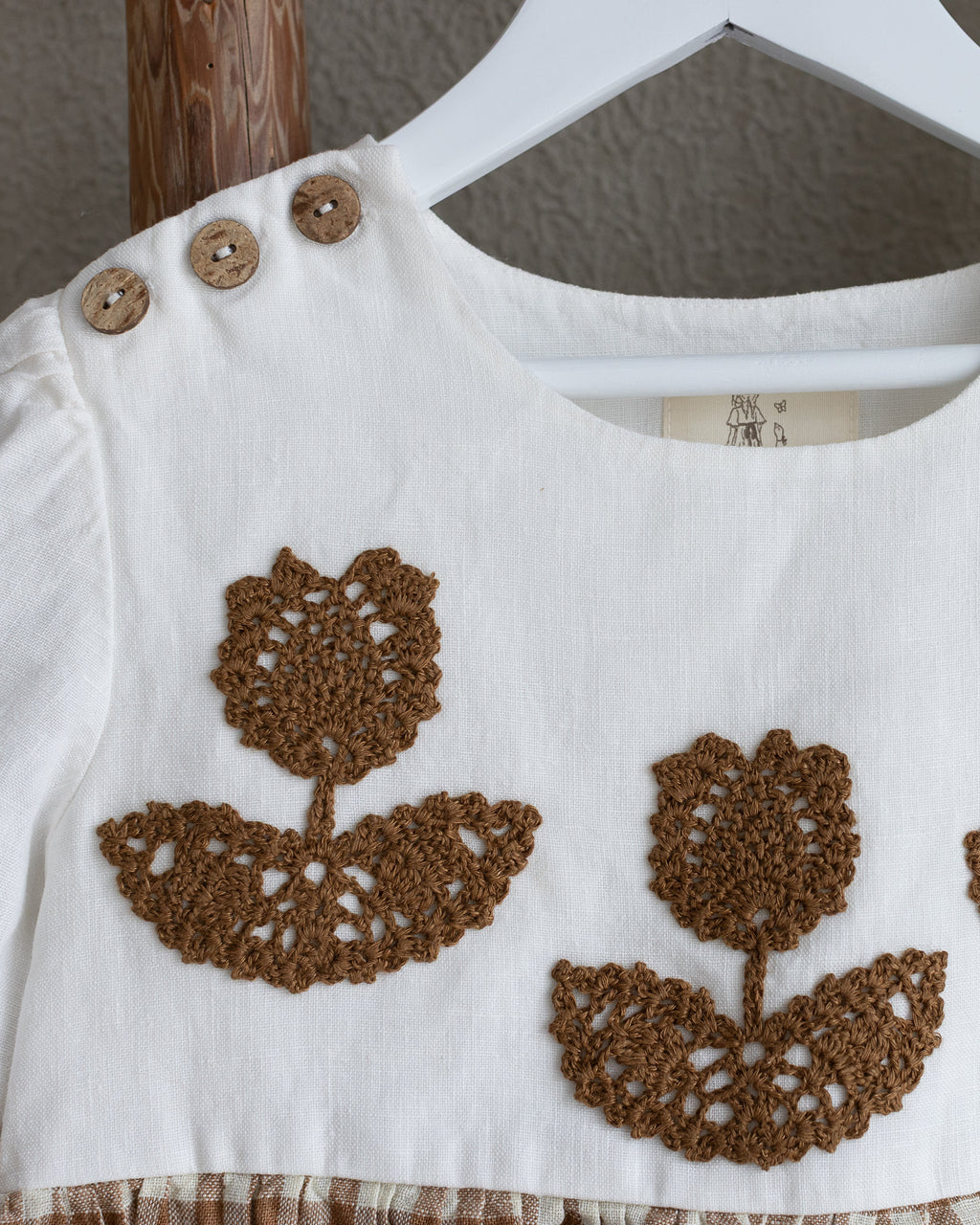 White linen dress top with brown crocheted flower on a hanger against a neutral background