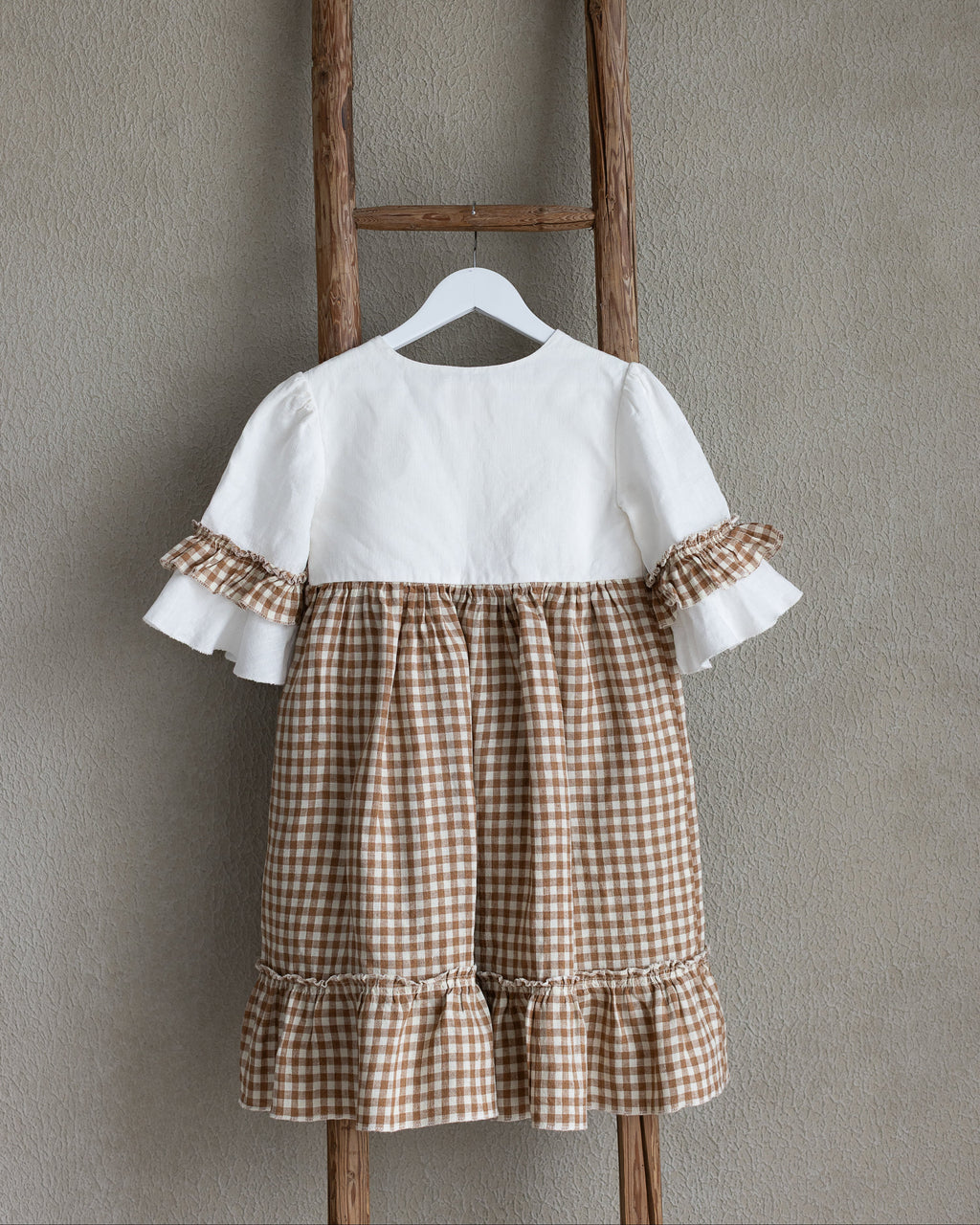 White linen blouse with brown checkered linen skirt hanging on a wooden ladder against a gray wall.