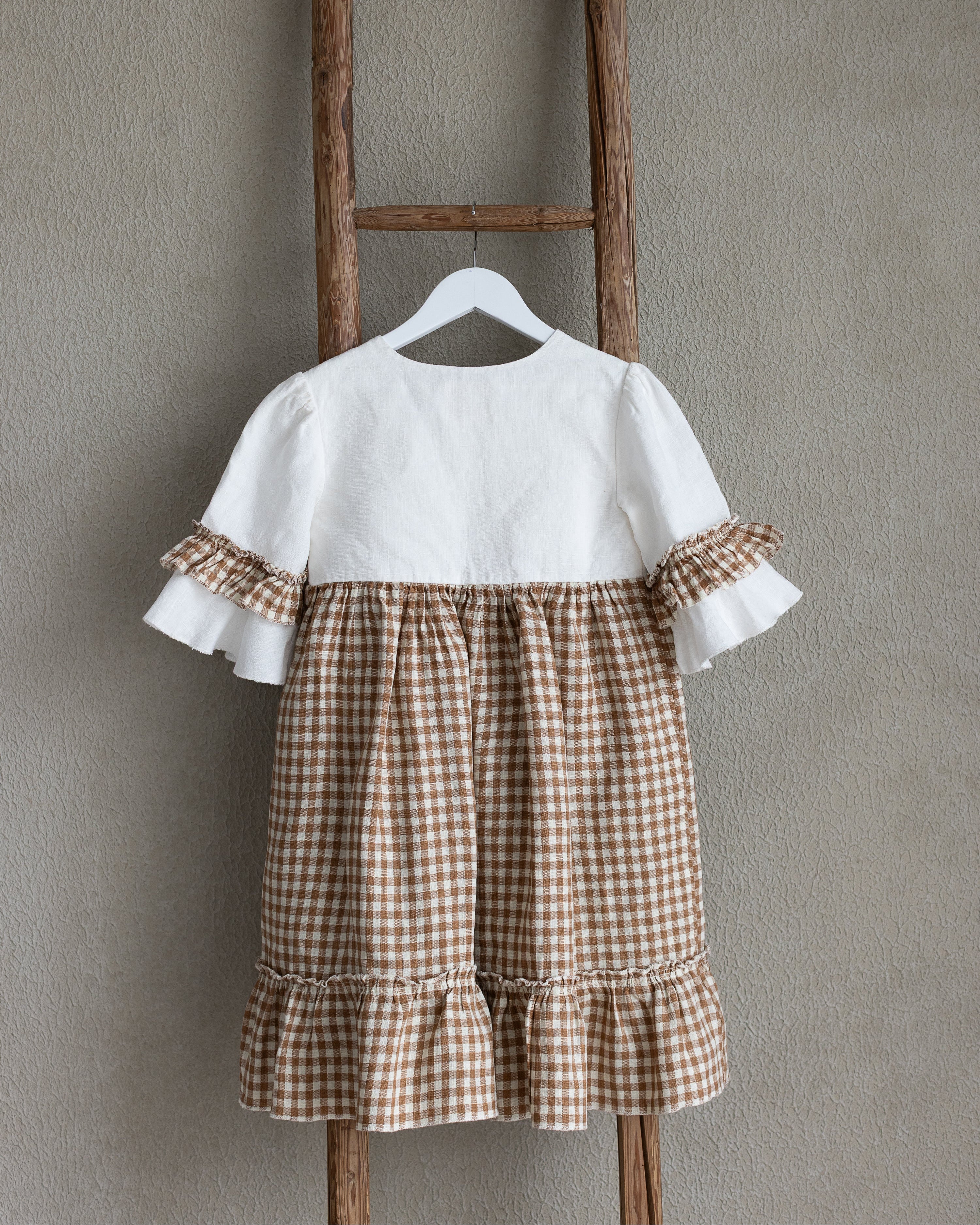 White linen blouse with brown checkered linen skirt hanging on a wooden ladder against a gray wall.
