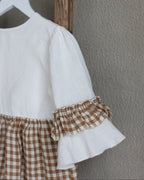 White linen blouse with ruffled sleeves and brown checkered linen skirt hanging on a wooden hanger against a gray wall.