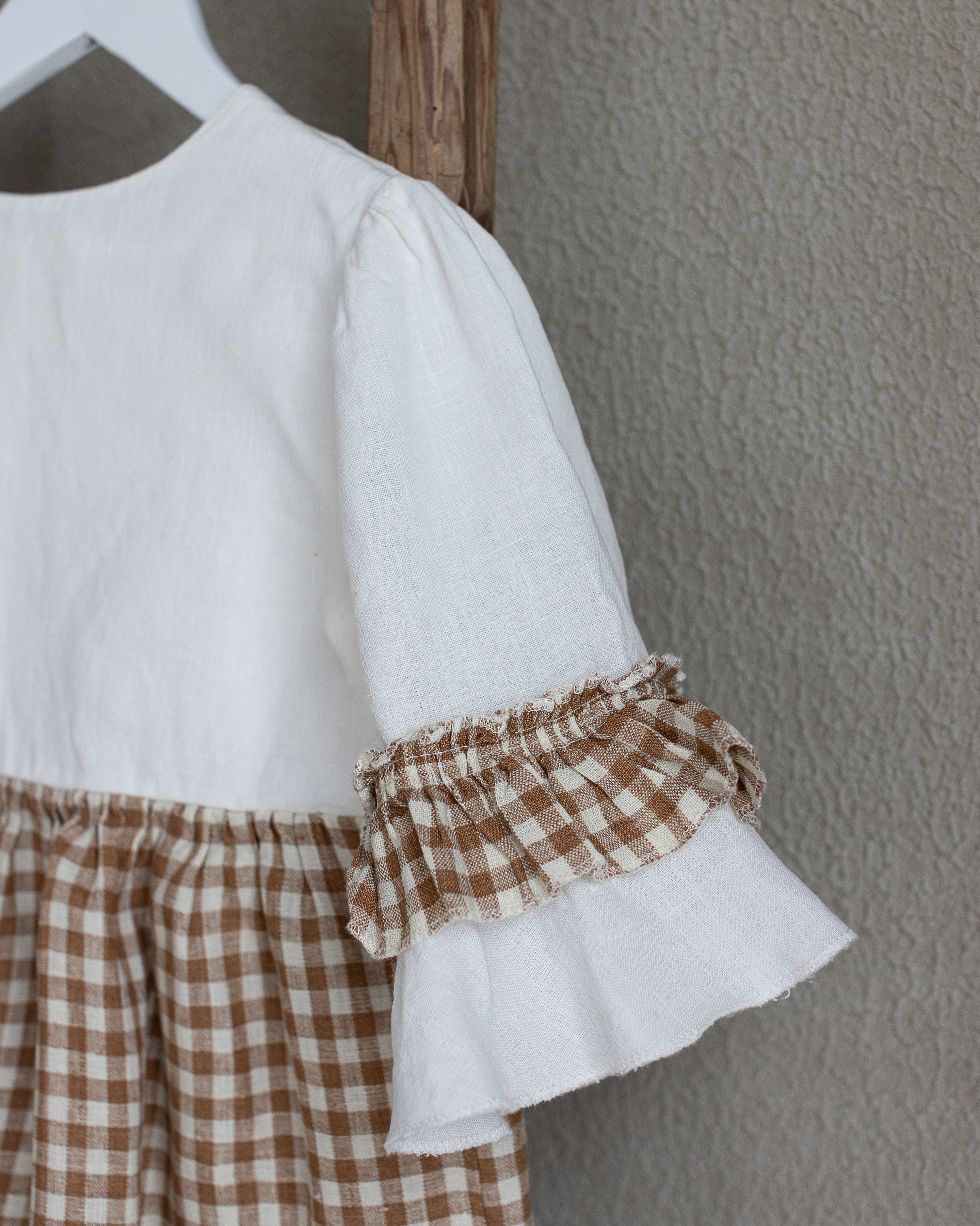 White linen blouse with ruffled sleeves and brown checkered linen skirt hanging on a wooden hanger against a gray wall.