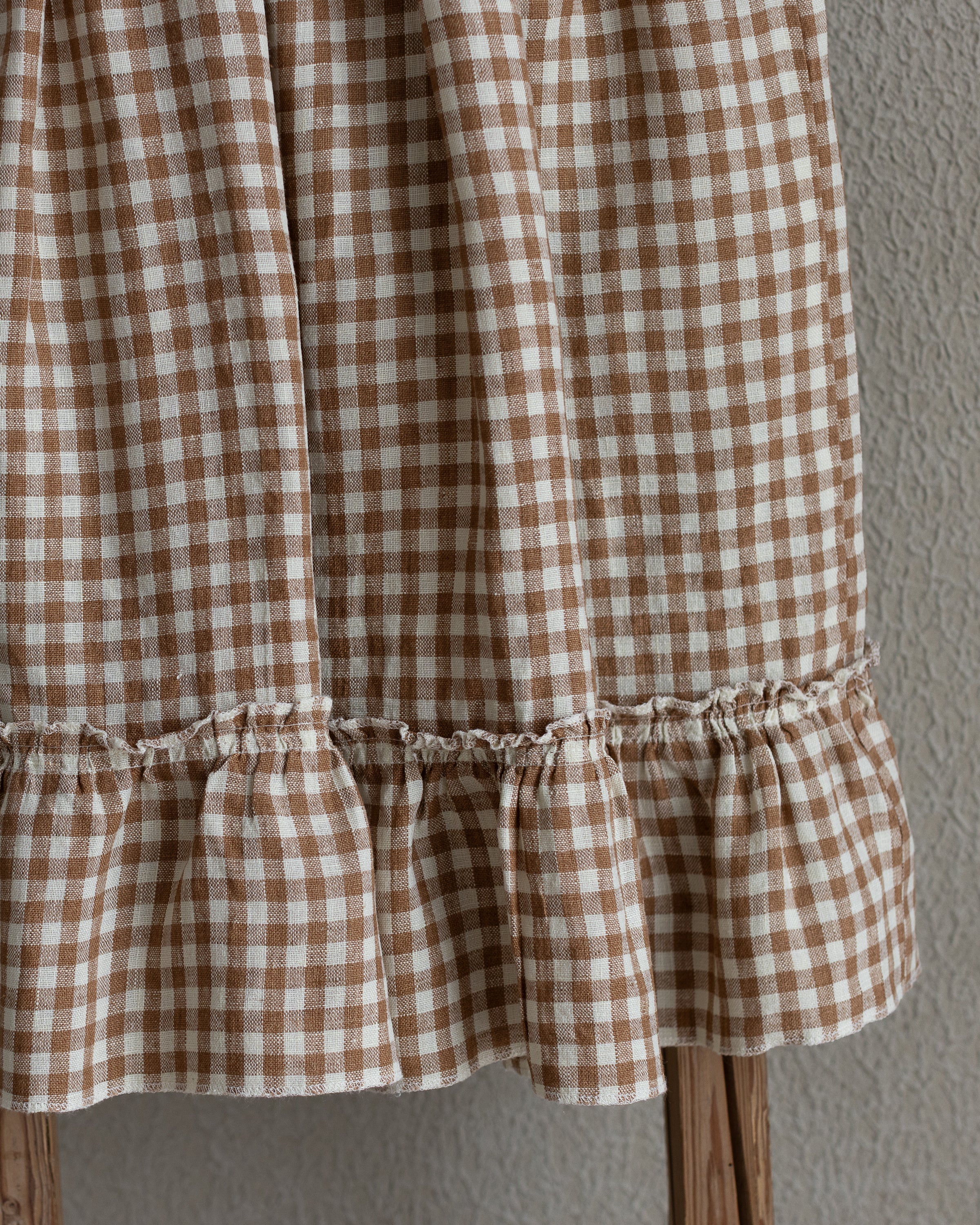 Brown and white checkered linen skirt with ruffle hem on a wooden stool against a gray wall.