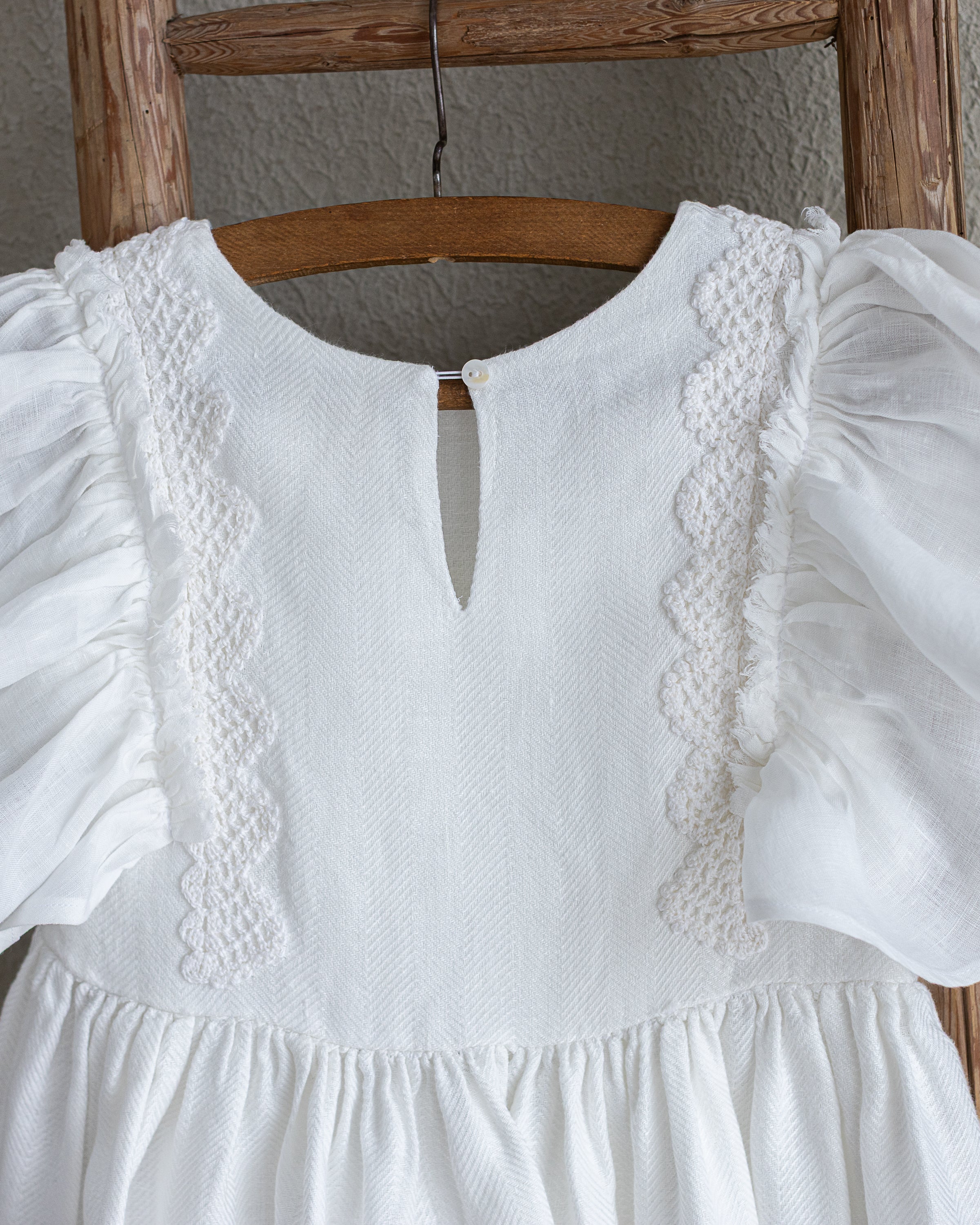 White linen dress with ruffled sleeves and crochet lace on a wooden ladder against a neutral wall.