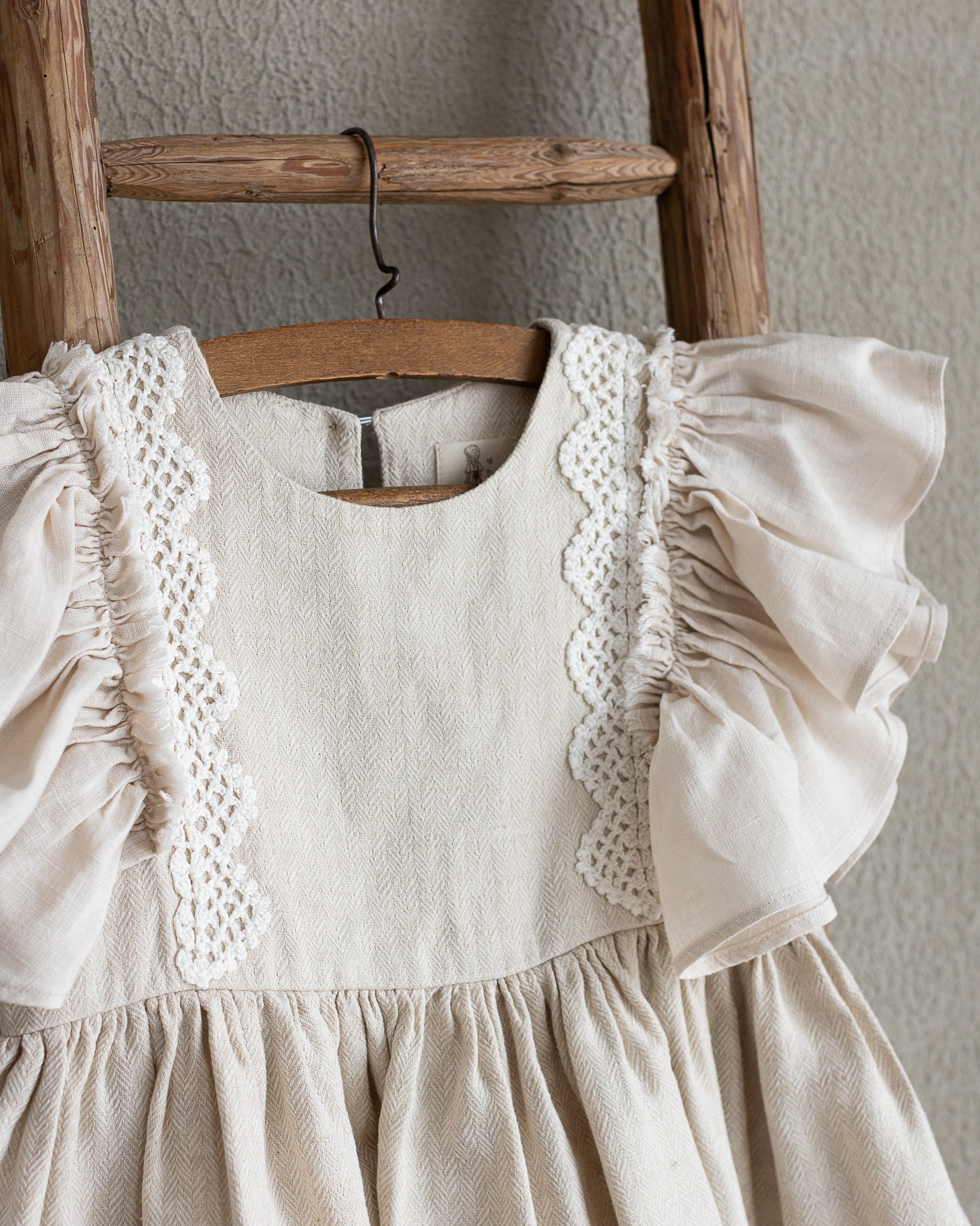 Beige linen dress with ruffled sleeves and crochet hanging on a wooden ladder against a neutral wall.