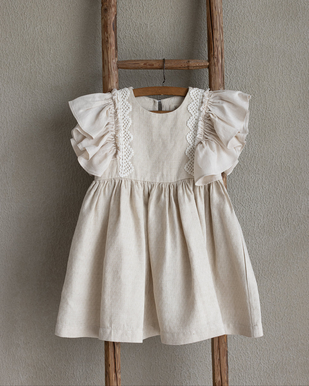 Beige linen dress for little girls with crochet lace details hanging on a wooden ladder against a plain wall.