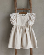 Beige linen dress for little girls with crochet lace details hanging on a wooden ladder against a plain wall.