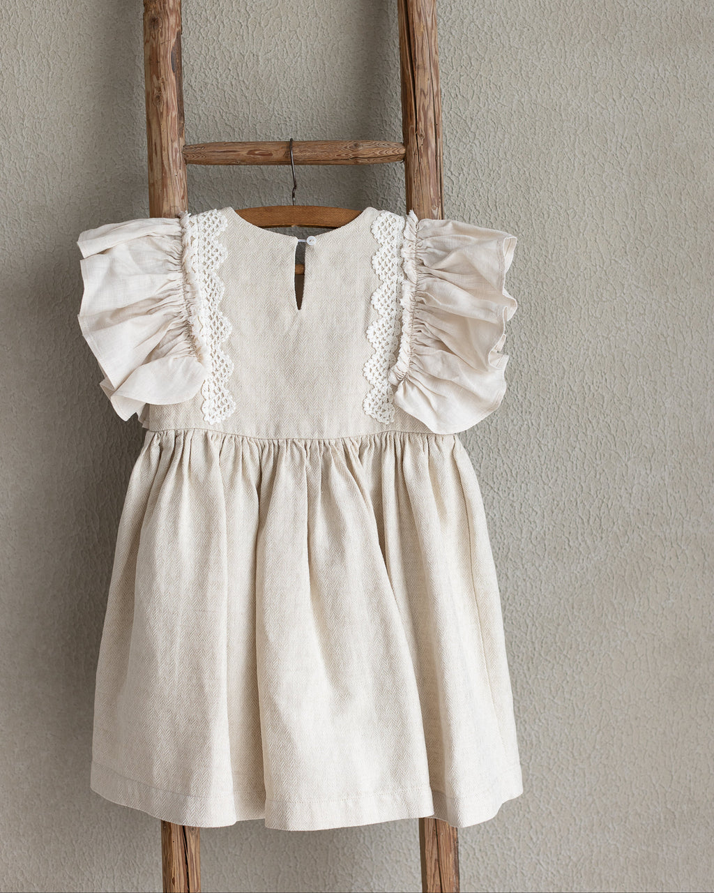 Light sand linen dress with ruffled sleeves and crocheted lace hanging on a wooden ladder against a beige wall.