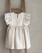 Light sand linen dress with ruffled sleeves and crocheted lace hanging on a wooden ladder against a beige wall.