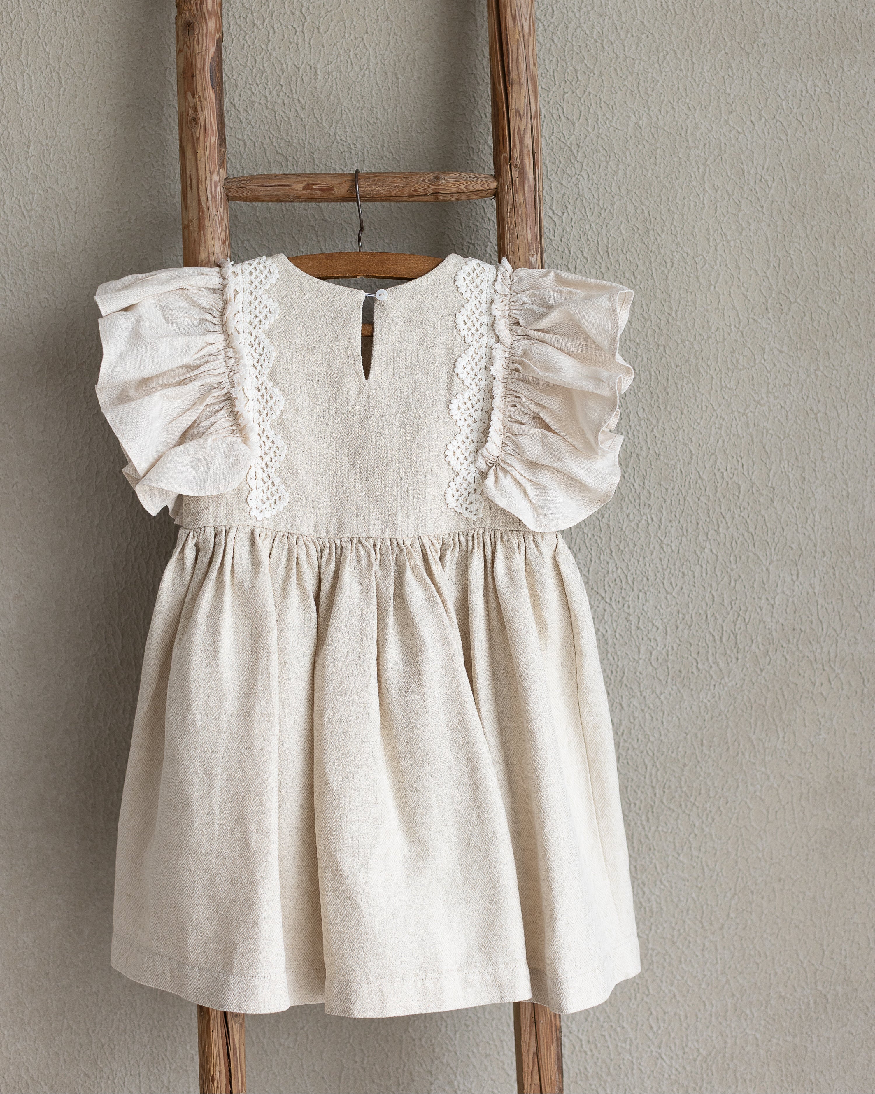 Light sand linen dress with ruffled sleeves and crocheted lace hanging on a wooden ladder against a beige wall.