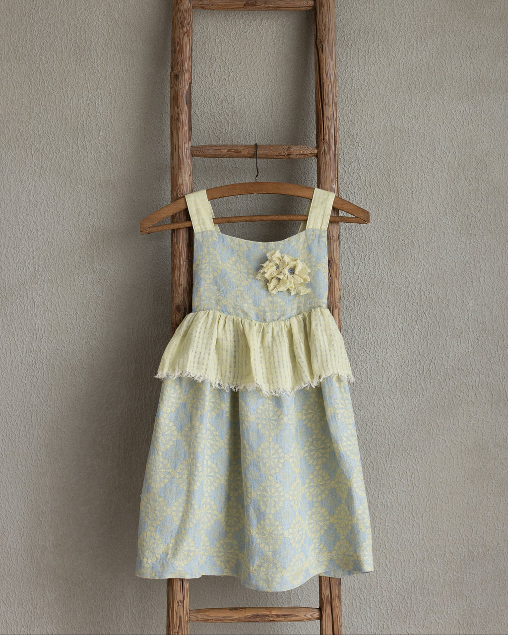 Toddler linen flolar dress with flower brooch hanging on a wooden ladder against a plain wall.