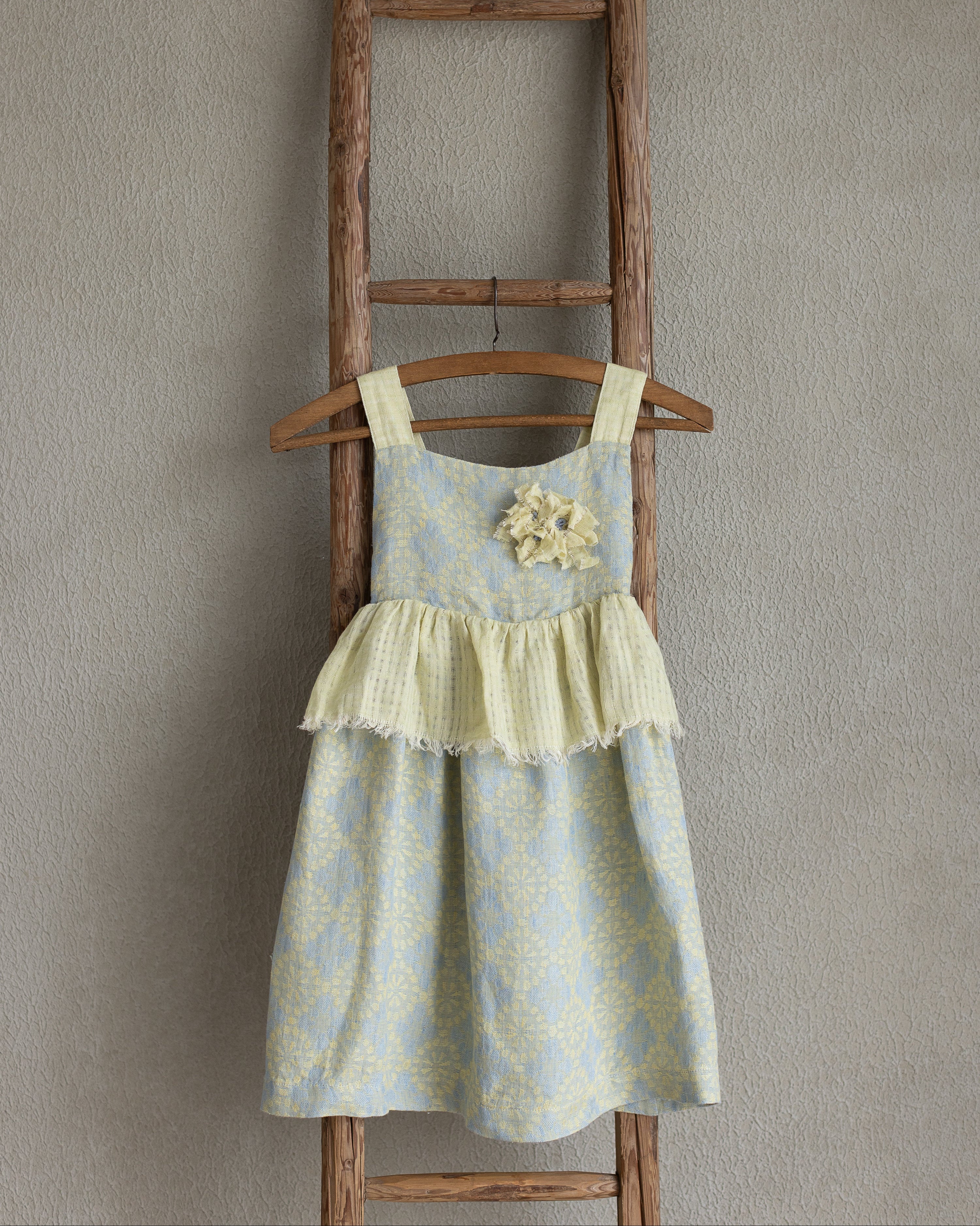 Toddler linen flolar dress with flower brooch hanging on a wooden ladder against a plain wall.