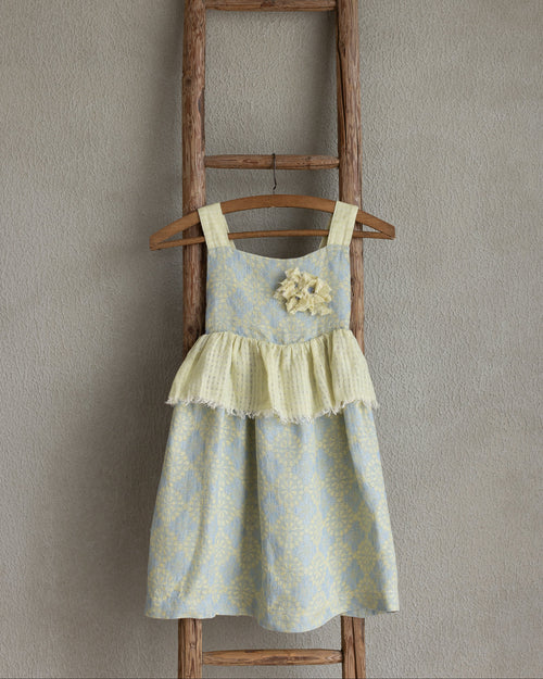 Toddler linen flolar dress with flower brooch hanging on a wooden ladder against a plain wall.
