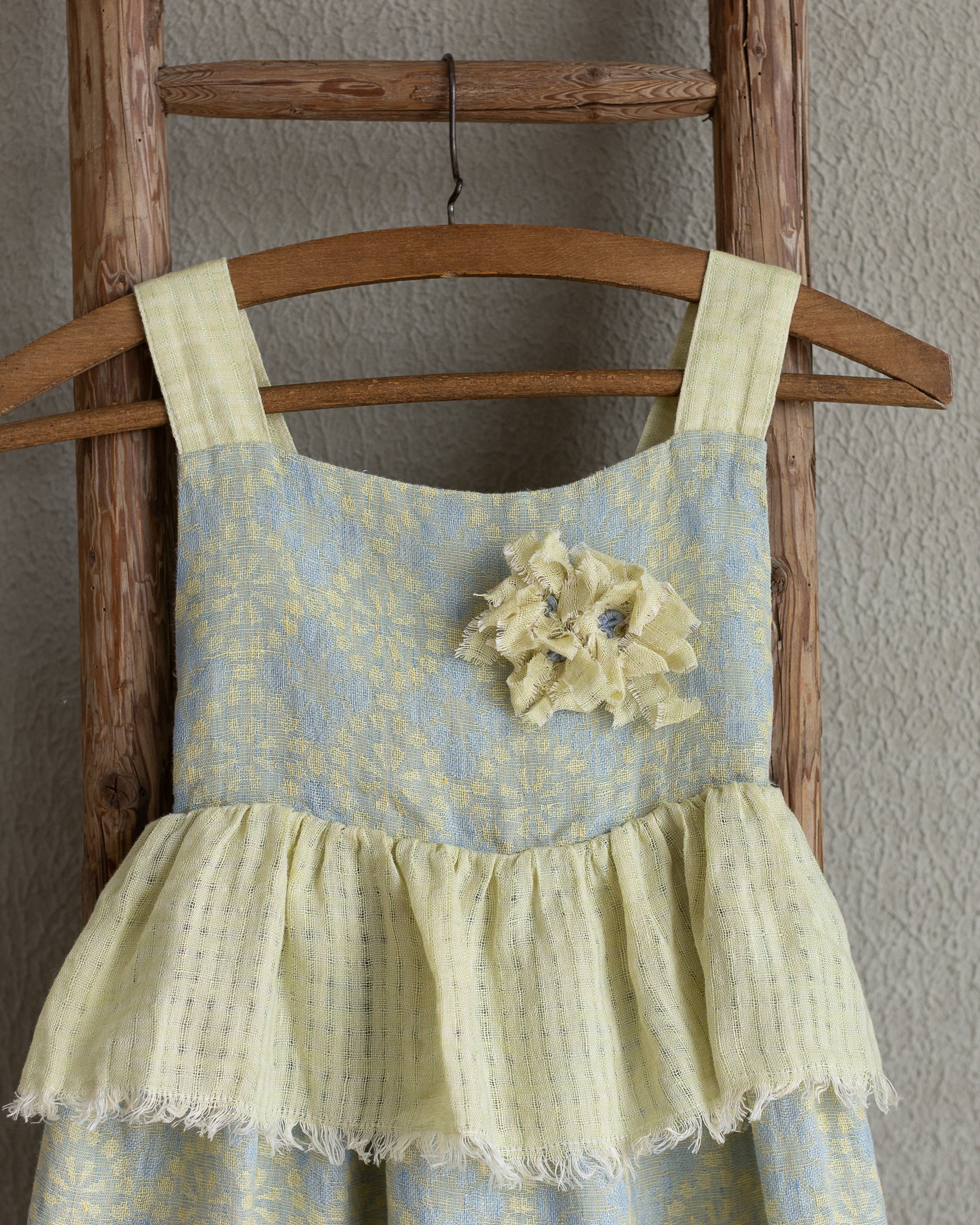 Girls linen flolar dress with flower brooch hanging on a wooden ladder against a plain wall.