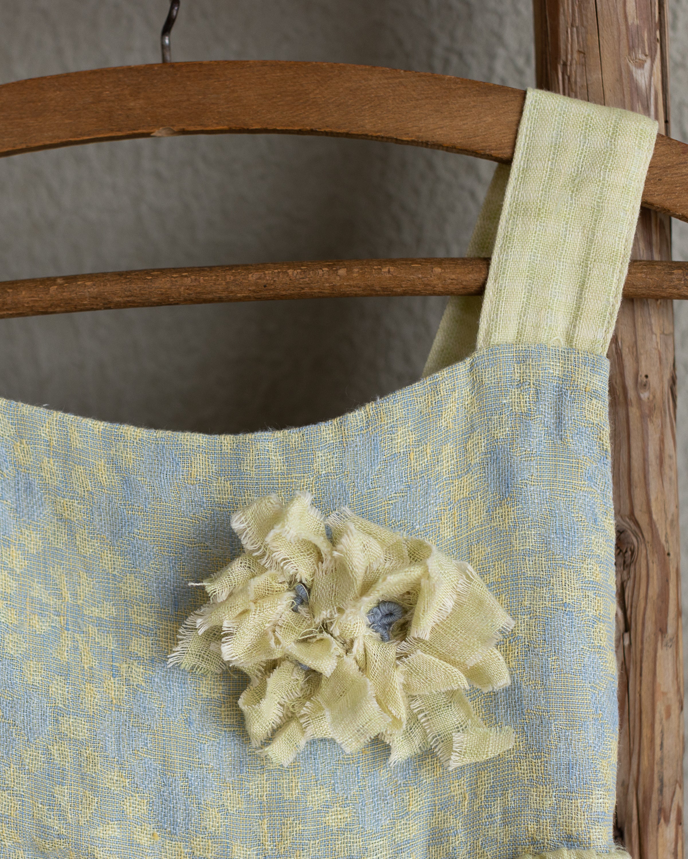 Linen floral dress for baby girl with flower decoration hanging on a wooden frame against a neutral background