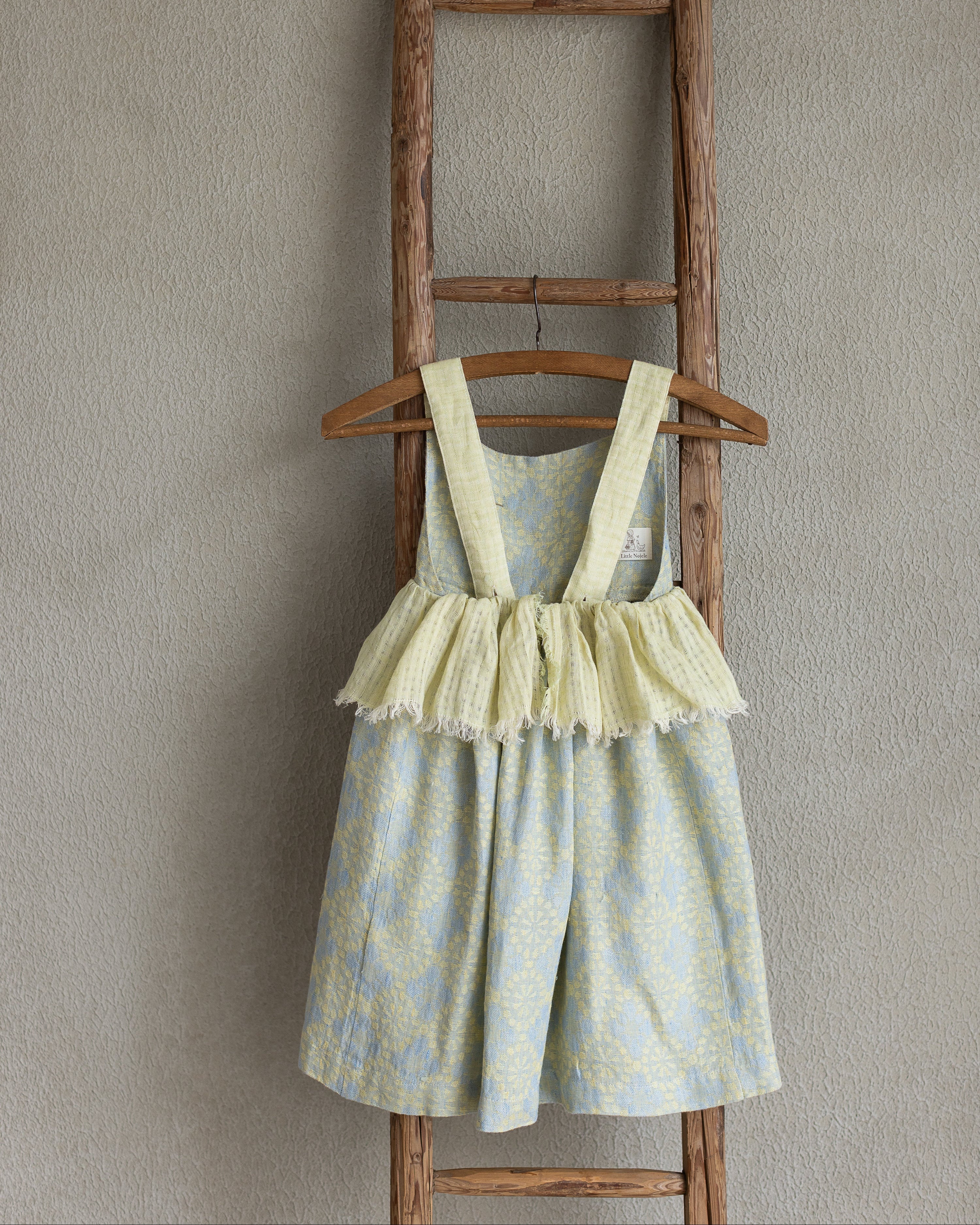 Girl's floral linen dress with ruffled skirt on a wooden hanger against a beige wall.