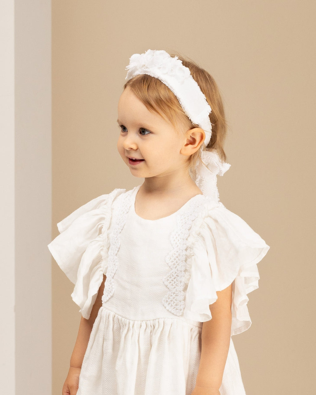 Young girl wearing a white linene dress with ruffled sleeves and a matching linen headband against a beige background