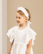 Young girl wearing a white linene dress with ruffled sleeves and a matching linen headband against a beige background