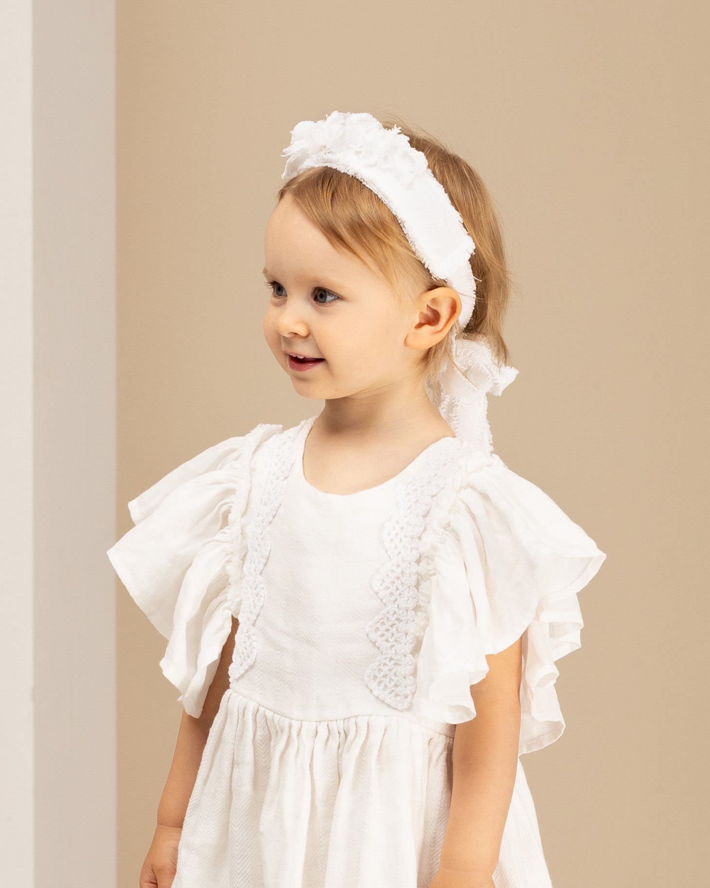Young girl wearing a white linene dress with ruffled sleeves and a matching linen headband against a beige background