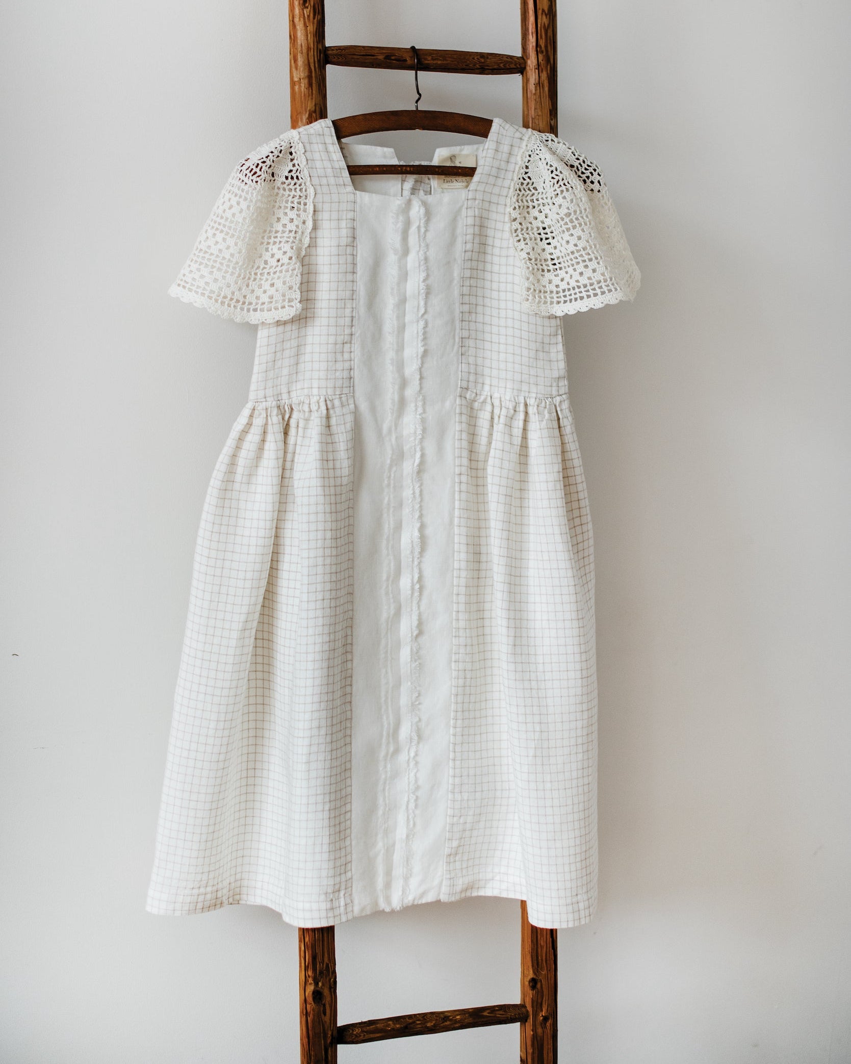 White checkered linen dress with crocheted sleeves on a wooden ladder against a white wall