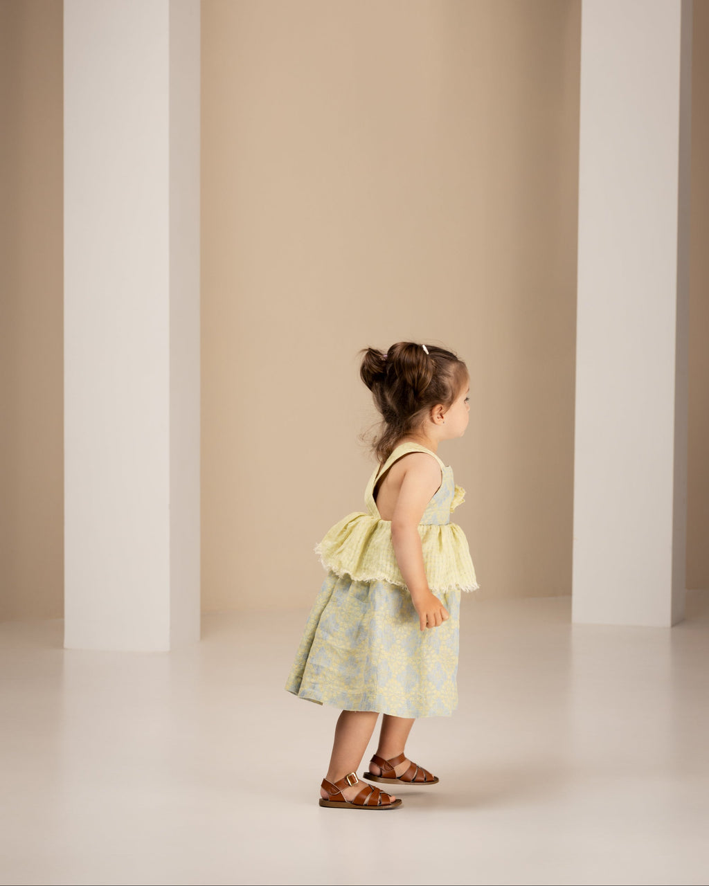 Little girl in a yellow floral linen dress standing in a studio with beige walls.