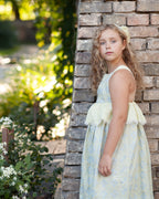 Young girl in a light linen dress standing against a brick wall with greenery in the background