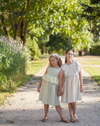 Two young girls in light linen dresses standing on a gravel path with greenery around.