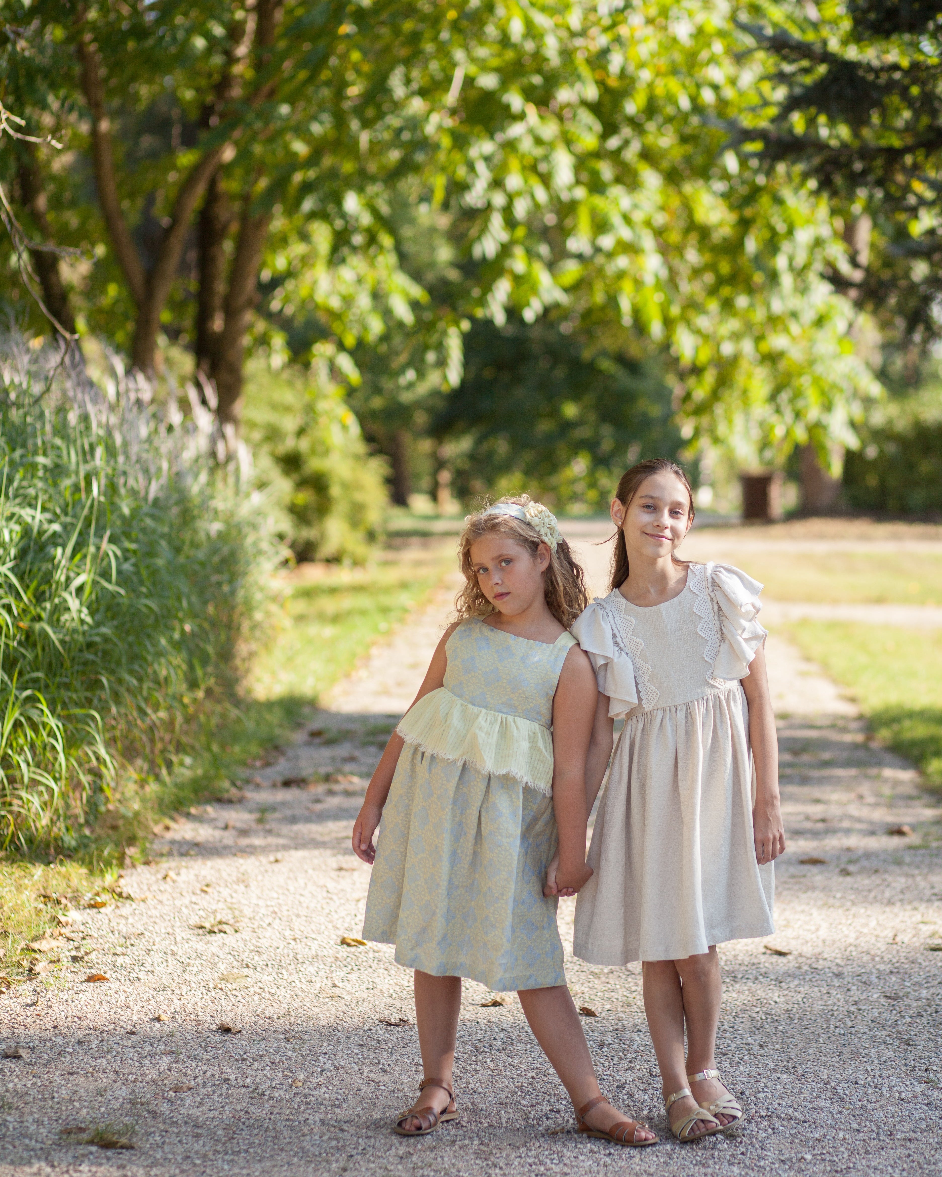 Two young girls in light linen dresses standing on a gravel path with greenery around.