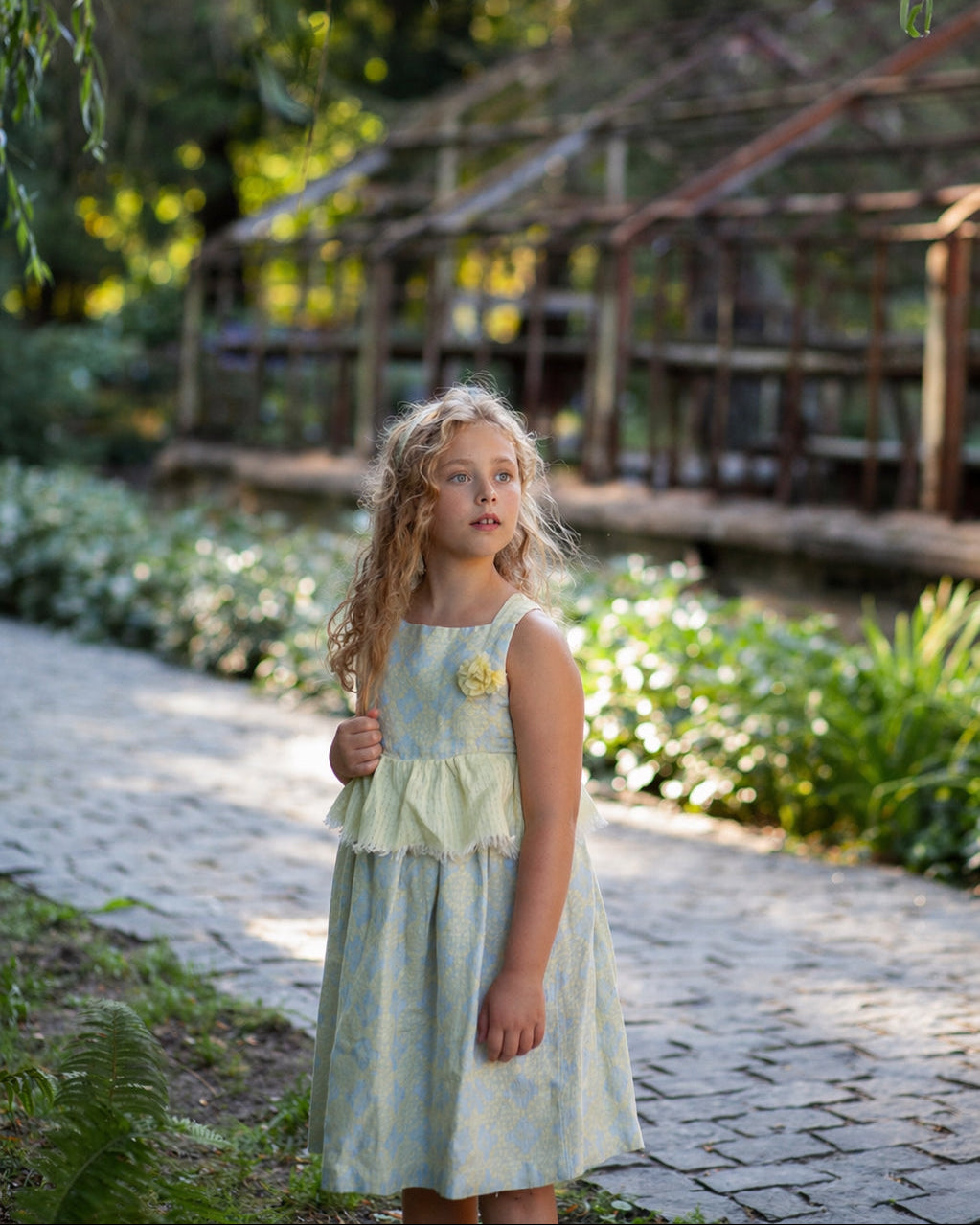Young girl in a light floral linen dress standing in nature and a wooden structure in the background.