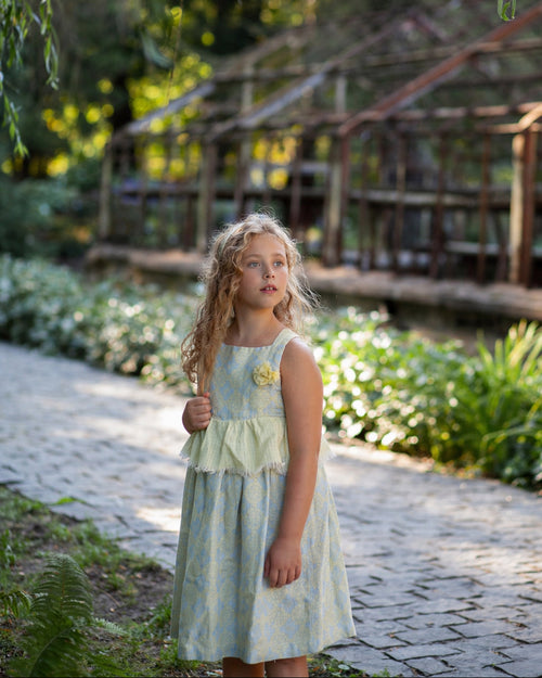 Young girl in a light floral linen dress standing in nature and a wooden structure in the background.