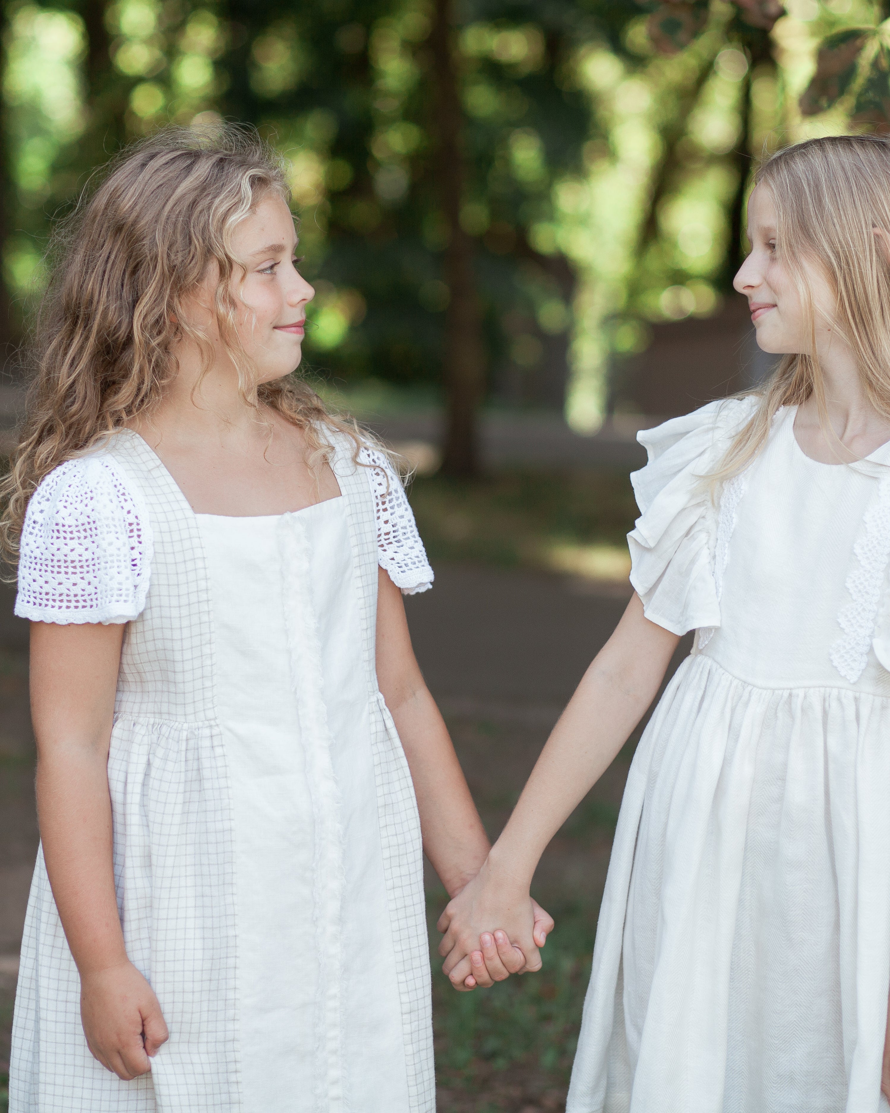 Two young girls in white linen dresses with crochet details holding hands outdoors with greenery in the background