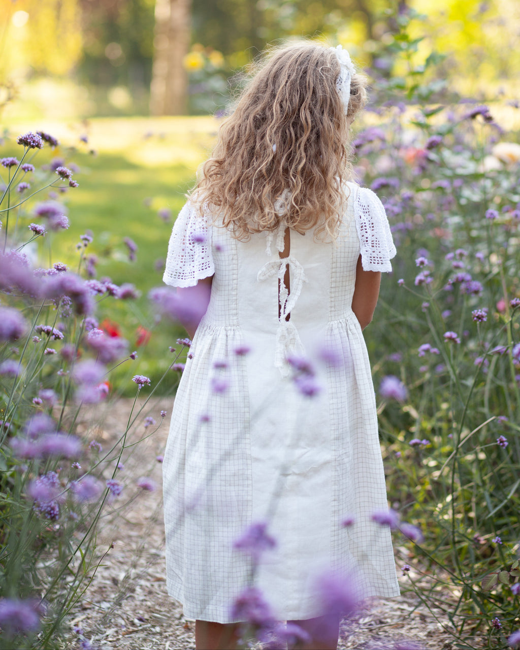 Young girl in a white linen dress with crocheted sleeves standing in a field of purple flowers with a blurred natural background