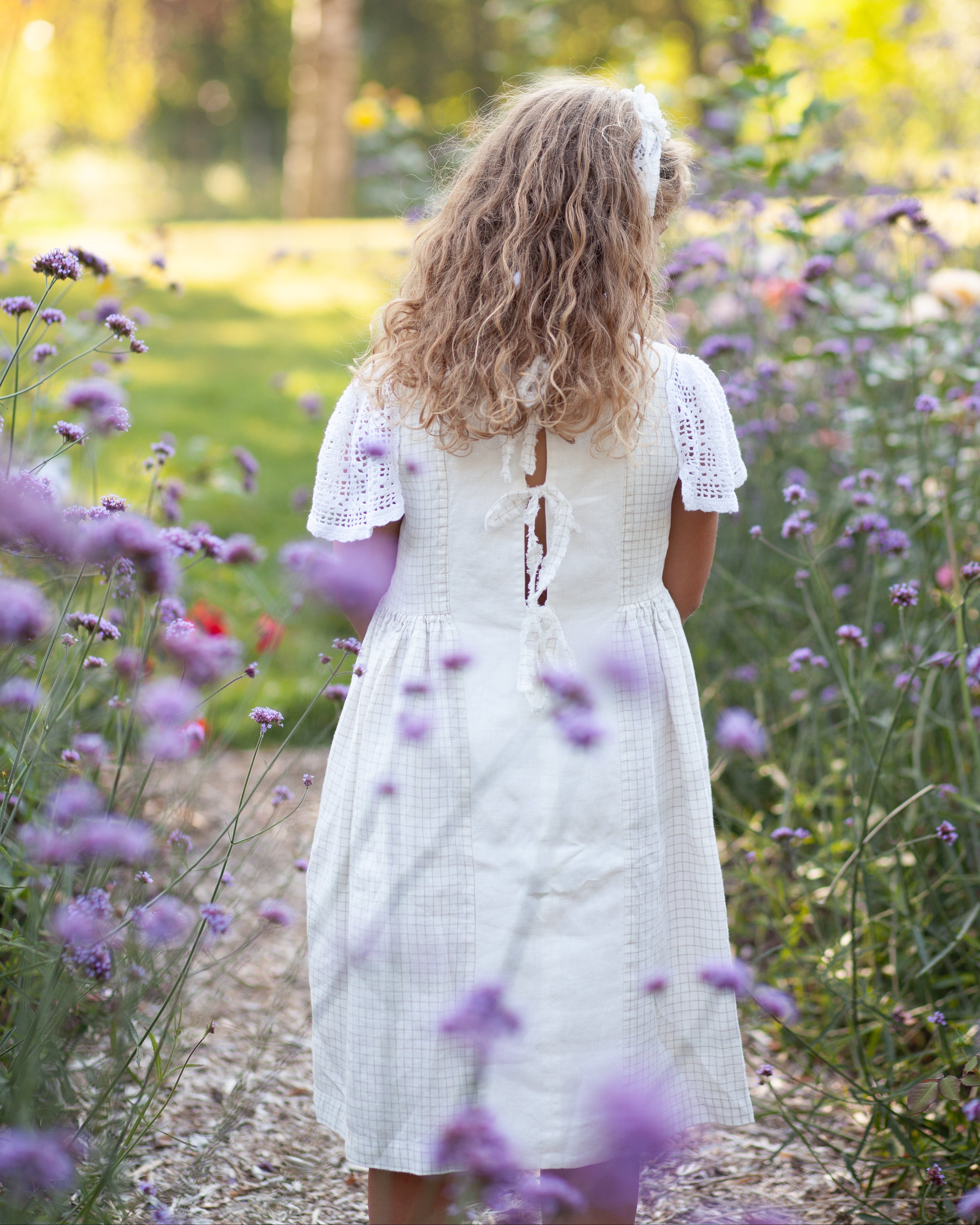 Young girl in a white linen dress with crocheted sleeves standing in a field of purple flowers with a blurred natural background