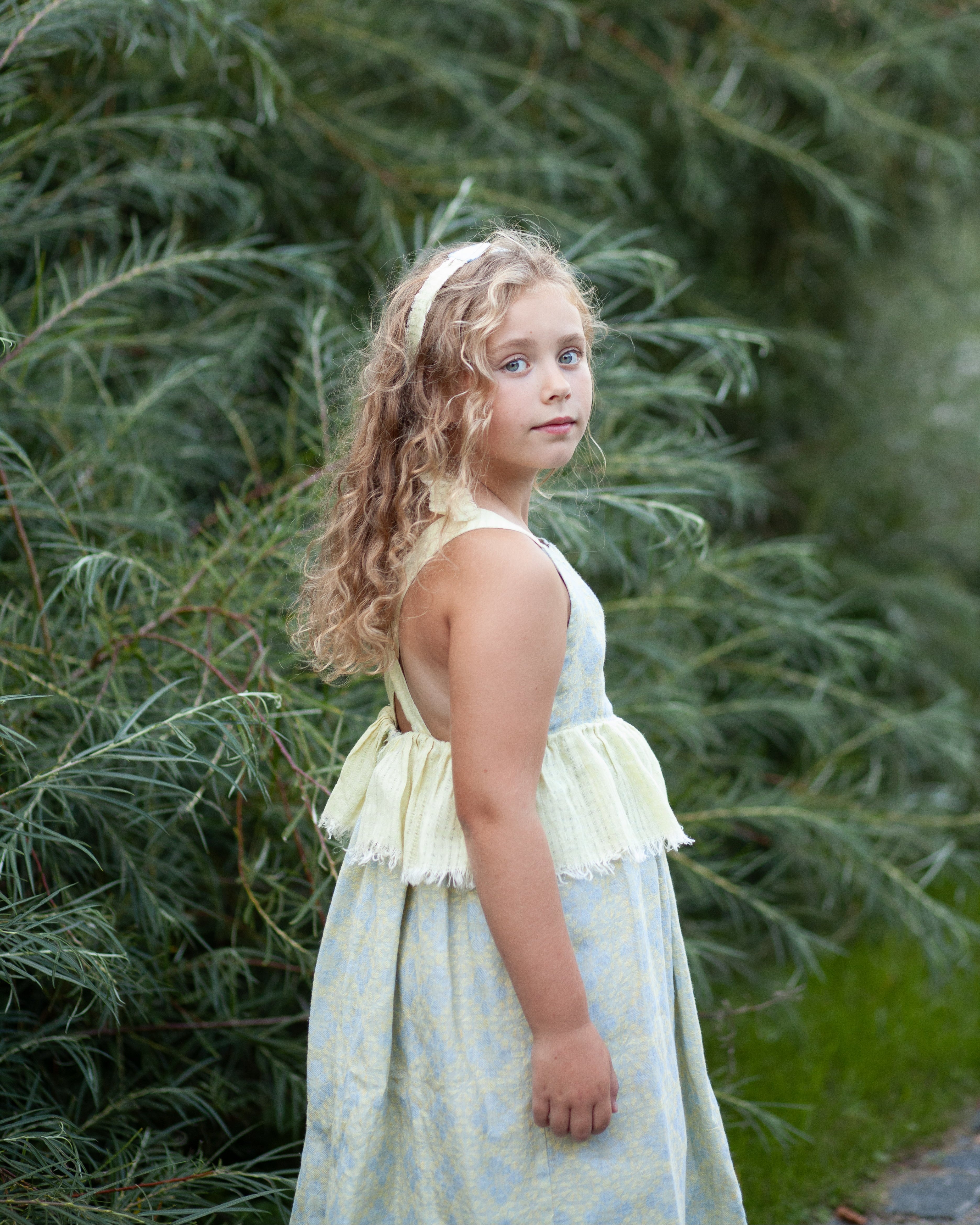 Young girl in a floral linen dress standing in front of green foliage