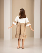 Little girl wearing linen dress with white top and brown checkered skirt standing in an archway.