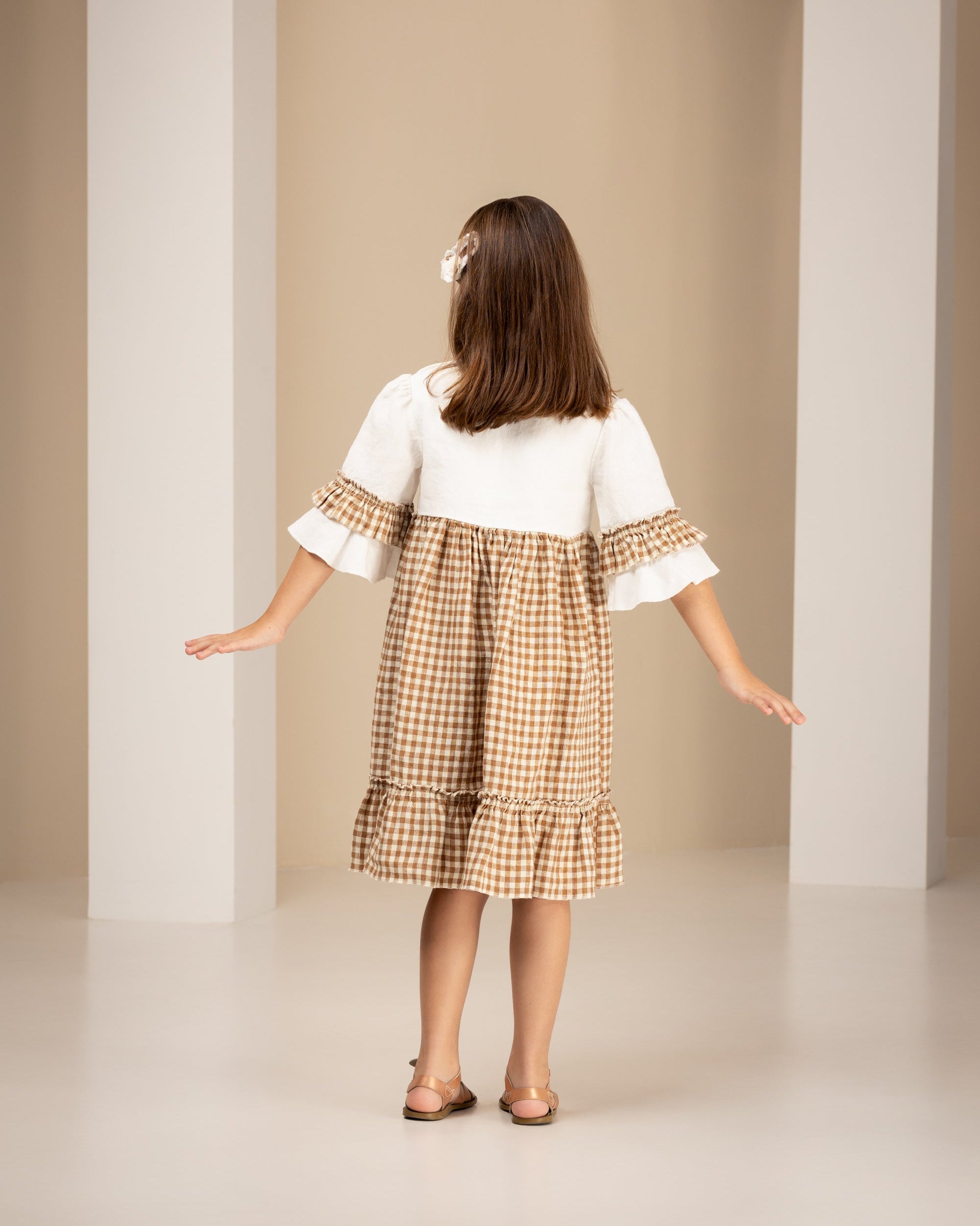 Little girl wearing linen dress with white top and brown checkered skirt standing in an archway.