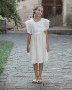 White linen dress with crochet flowers, Romantic heirloom clothing for girl, White special occasion outfit, Baptism dress for toddler, A young girl with braided hair stands on a cobblestone path wearing a white linen short-sleeve dress with ruffled shoulders and crochet flowers on the bodice, paired with metallic sandals.