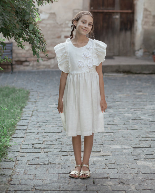 White linen dress with crochet flowers, Romantic heirloom clothing for girl, White special occasion outfit, Baptism dress for toddler, A young girl with braided hair stands on a cobblestone path wearing a white linen short-sleeve dress with ruffled shoulders and crochet flowers on the bodice, paired with metallic sandals.