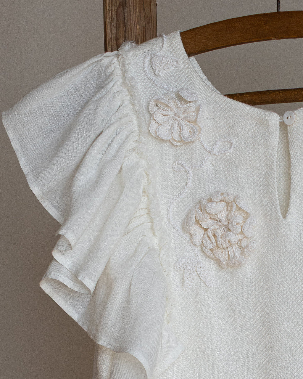 White linen dress with crochet flowers, Romantic heirloom clothing for girl, White special occasion outfit, Baptism dress for toddler, An white linen dress with ruffled sleeves and crochet flowers, hanging on a wooden hanger against a neutral background.