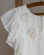 White linen dress with crochet flowers, Romantic heirloom clothing for girl, White special occasion outfit, Baptism dress for toddler, An white linen dress with ruffled sleeves and crochet flowers, hanging on a wooden hanger against a neutral background.