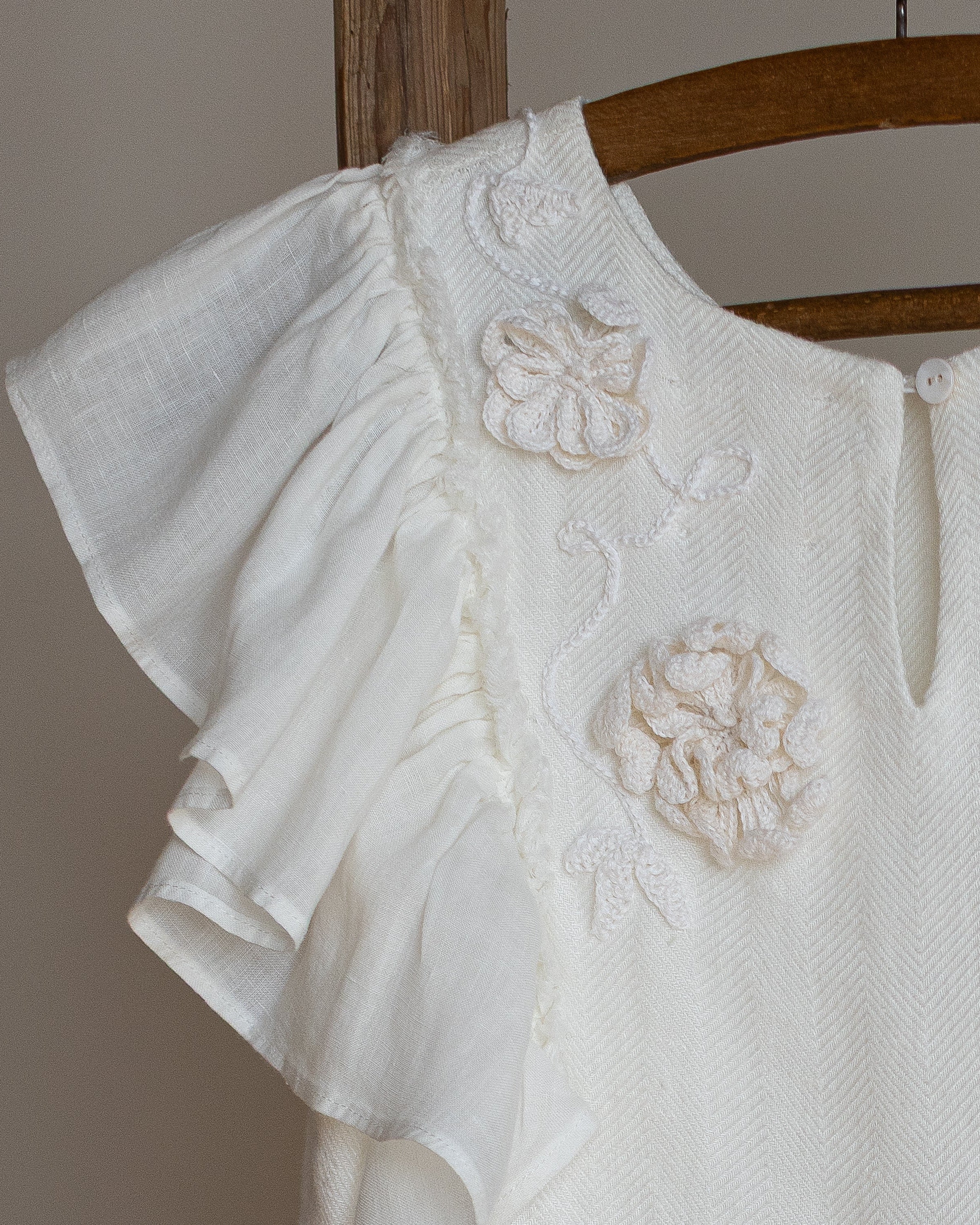 White linen dress with crochet flowers, Romantic heirloom clothing for girl, White special occasion outfit, Baptism dress for toddler, An white linen dress with ruffled sleeves and crochet flowers, hanging on a wooden hanger against a neutral background.