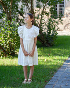 White linen dress with crochet flowers, Romantic heirloom clothing for girl, White special occasion outfit, Baptism dress for toddler, A young girl stands outdoors on a grassy lawn wearing a white linen short-sleeved dress with ruffled sleeves and a floral crochet details on the chest. The background features green trees, grass, and a stone pathway.