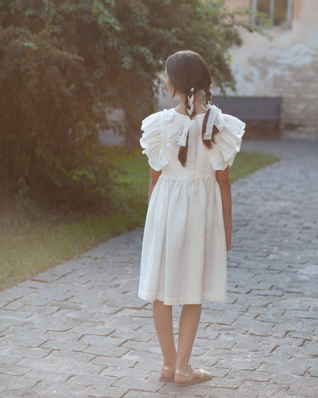 White linen dress with crochet flowers, Romantic heirloom clothing for girl, White special occasion outfit, Baptism dress for toddler, A young girl with braided hair stands on a stone path outdoors, viewed from behind, wearing a white linen dress with ruffled short sleeves and crochet flowers on the back.
