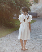 White linen dress with crochet flowers, Romantic heirloom clothing for girl, White special occasion outfit, Baptism dress for toddler, A young girl with braided hair stands on a stone path outdoors, viewed from behind, wearing a white linen dress with ruffled short sleeves and crochet flowers on the back.