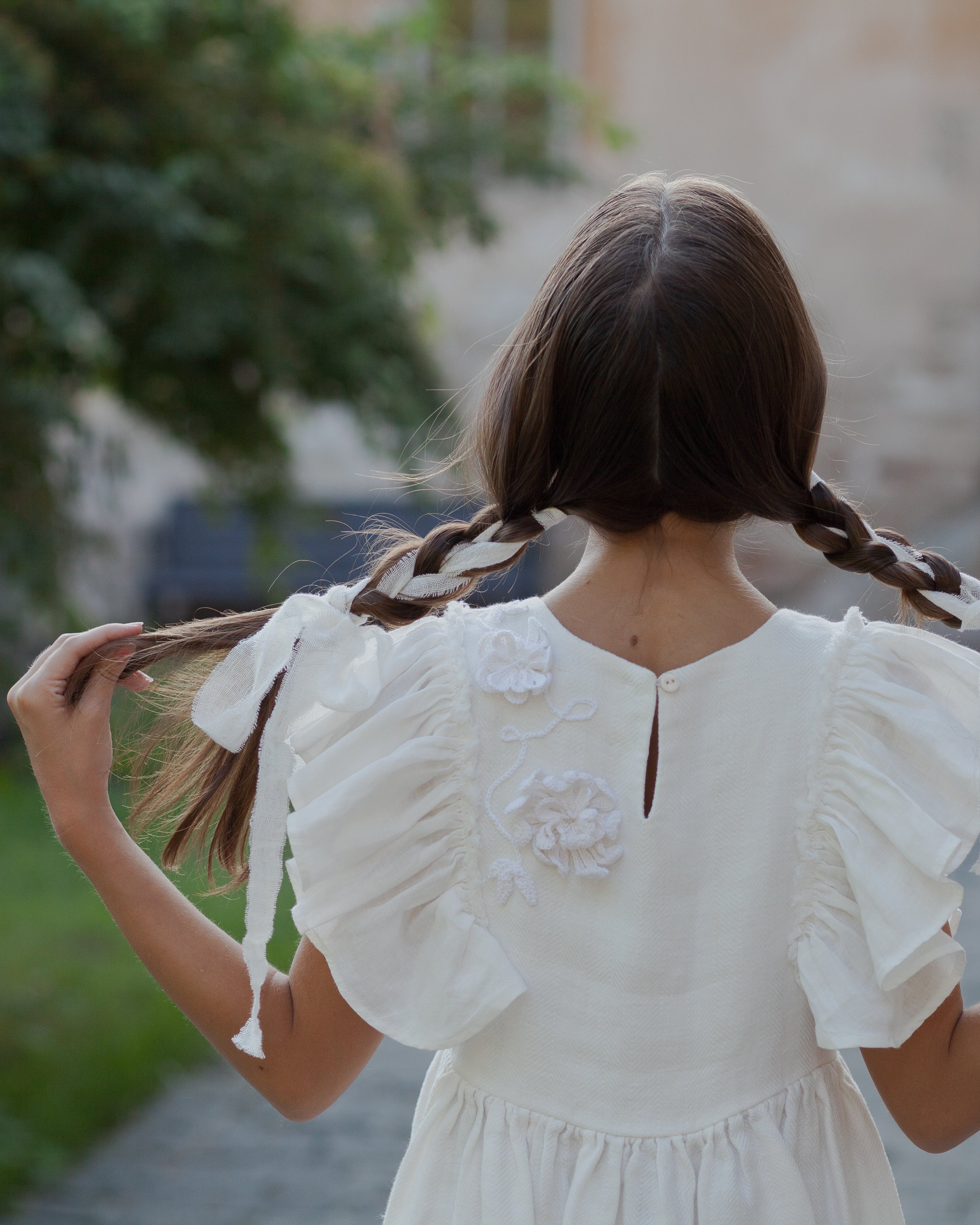 White linen dress with crochet flowers, Romantic heirloom clothing for girl, White special occasion outfit, Baptism dress for toddler, A young girl viewed from behind, wearing a white ruffled dress and holding up her long brown hair styled in two braids, set against a blurred outdoor background of greenery and a stone building.