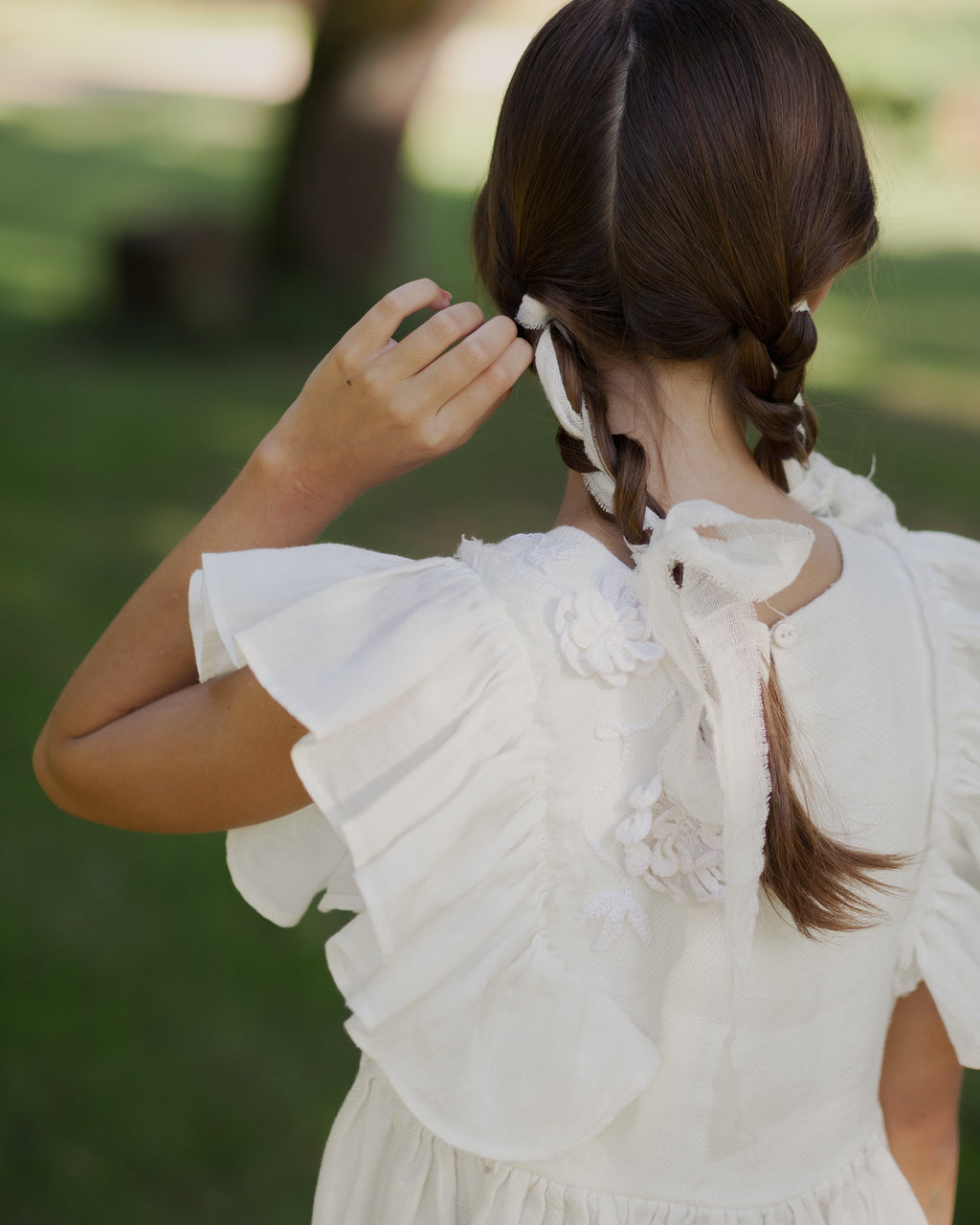 White linen dress with crochet flowers, Romantic heirloom clothing for girl, White special occasion outfit, Baptism dress for toddler, A young girl seen from behind wearing a white linen ruffled dress, adjusting a white hair tie with braided hair, in an outdoor park setting.
